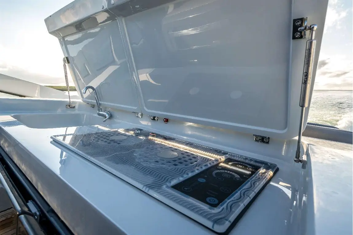 A sleek outdoor kitchen area on a yacht features a sink, controls, and a raised lid, with reflections of water in the background.